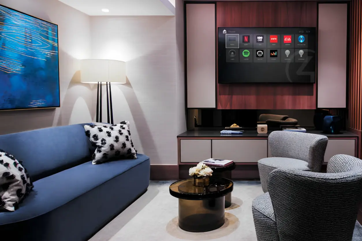 Modern living room with a blue sofa, abstract art, a lamp, two gray chairs, a small round table, and a wall-mounted TV displaying various app icons.