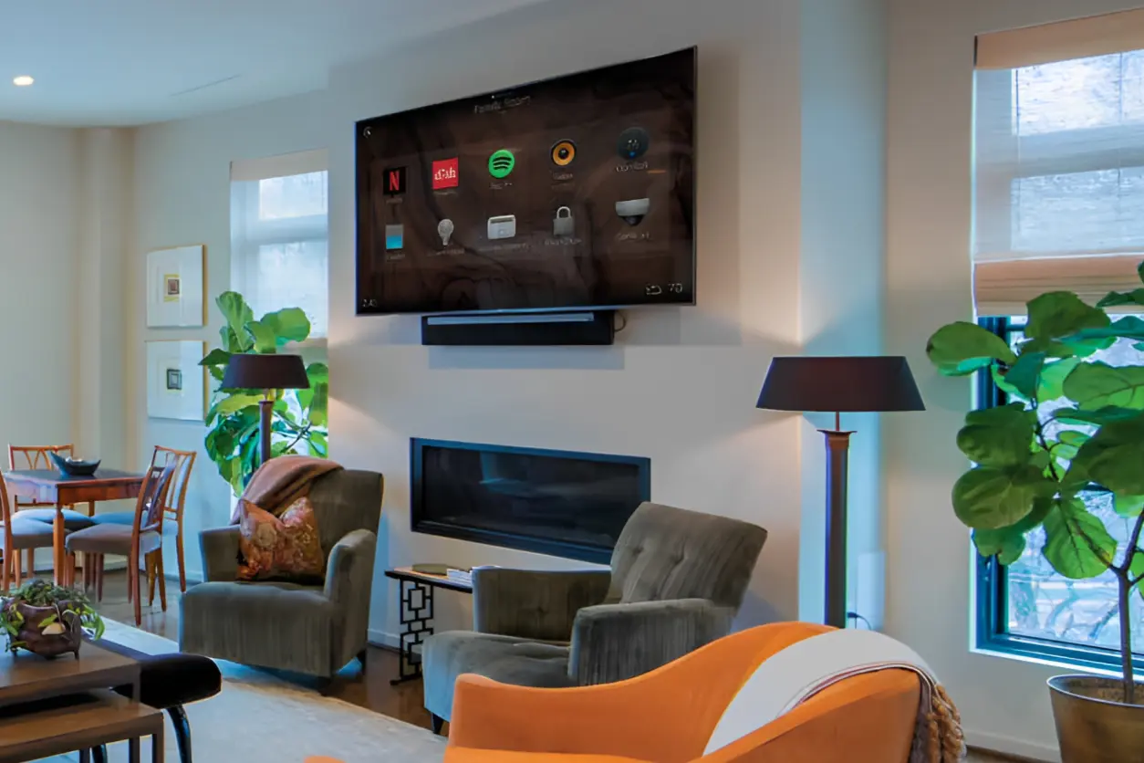 Living room with wall-mounted TV displaying streaming apps. Two armchairs face the TV, a fireplace below, and a dining area in the background. Large windows and indoor plants are present.