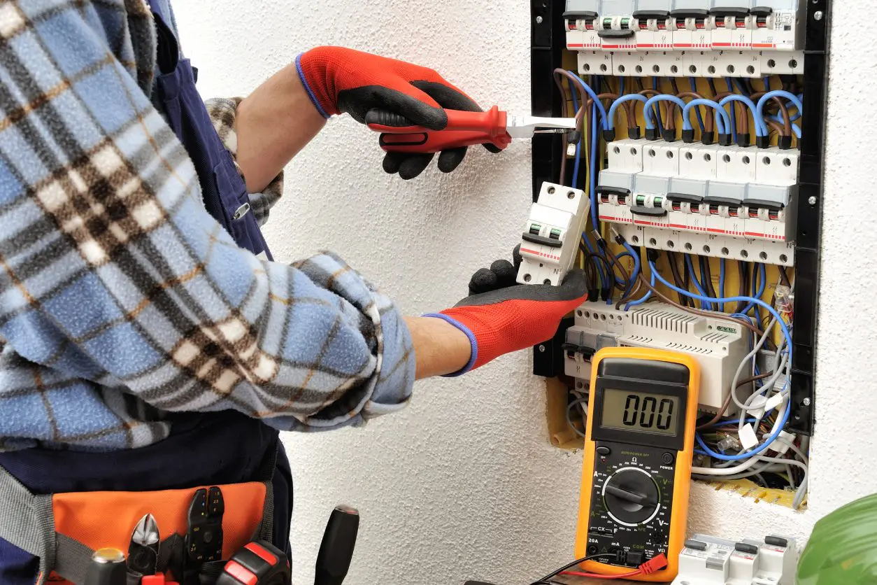 Electrician in plaid shirt and orange gloves uses a screwdriver on a circuit breaker panel with a multimeter showing 0.00 placed nearby.